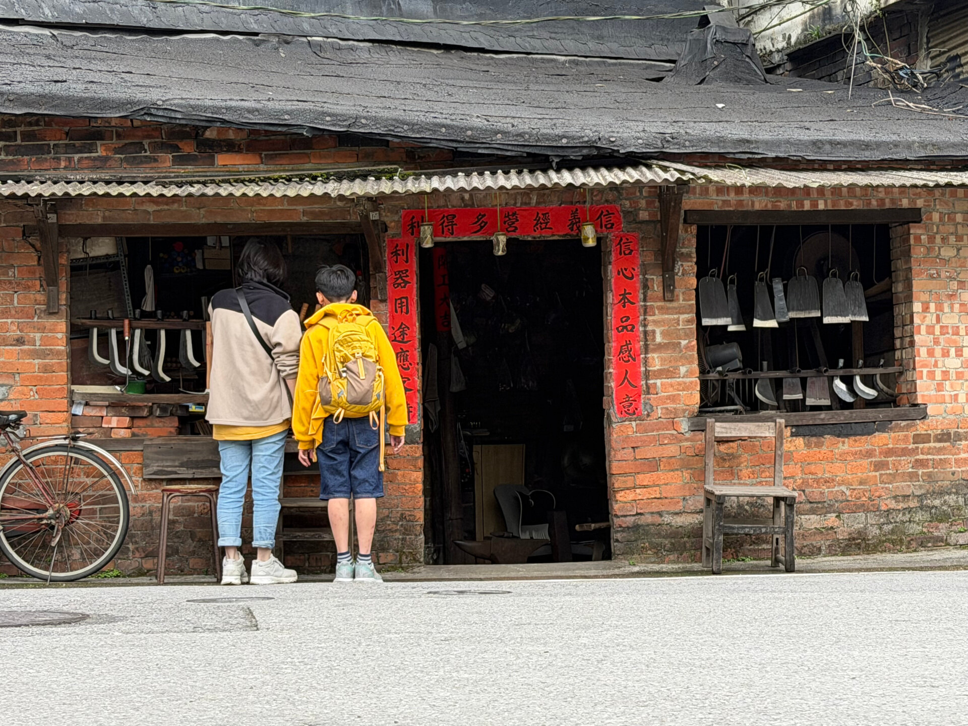 兩位行人駐足在一間傳統打鐵店前，紅色春聯上寫著「信義經營多得利、信心本品感人意」